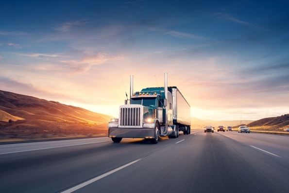 A teal colored 18 wheeler truck carrying cargo driving on a highway with multiple cards trailing behind it for Truck & Cargo Insurance.